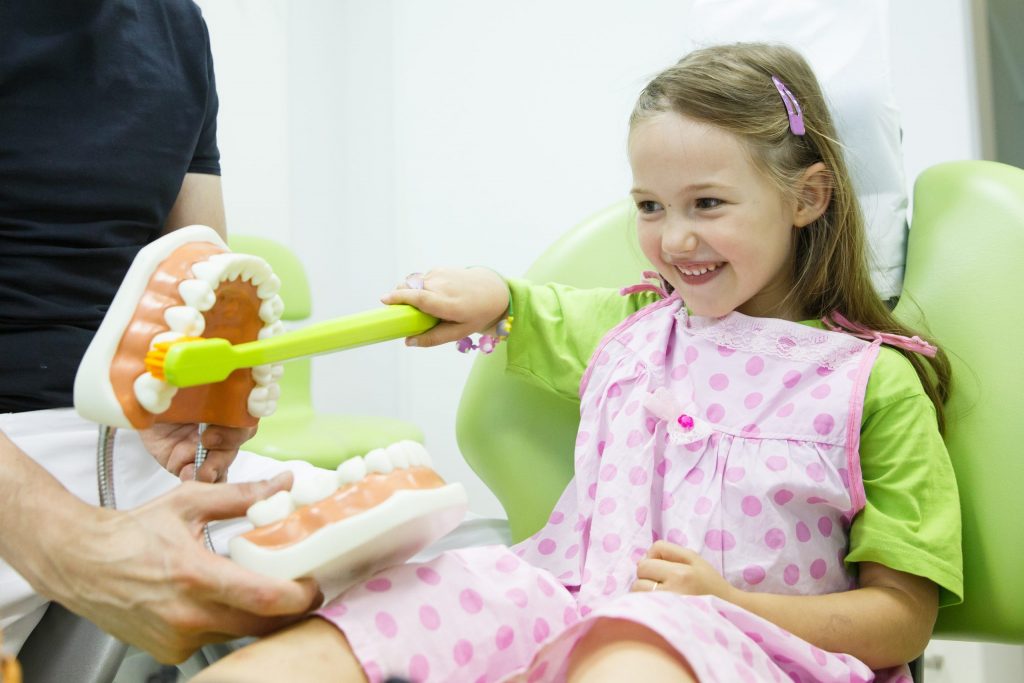 A small girl brushes a large model mouth with a big toothbrush. She's smiling during her pediatric dentistry visit.